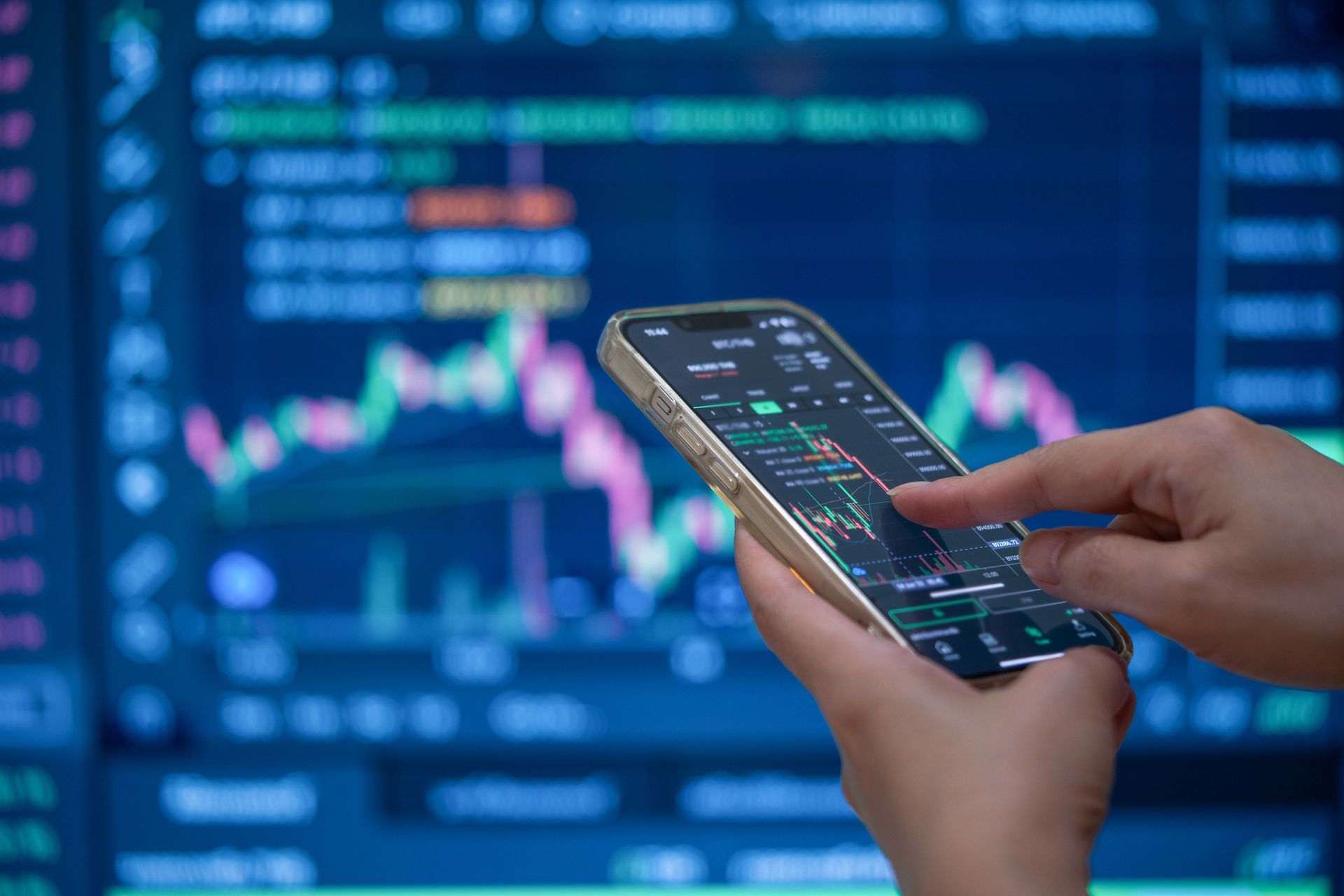 Person using a smartphone to view stock charts, with a large computer monitor displaying similar data in the background.
