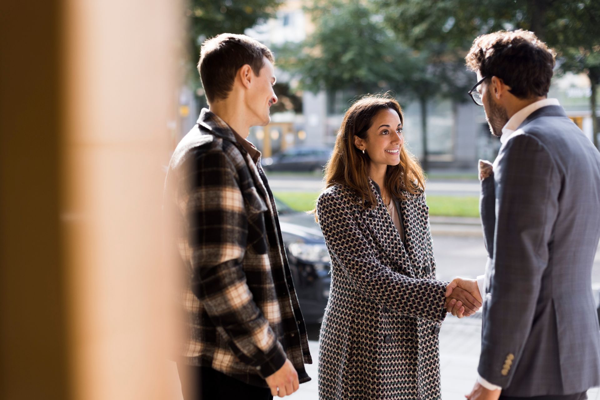 Three people greeting outdoors: woman shaking hands with man, another man watching.