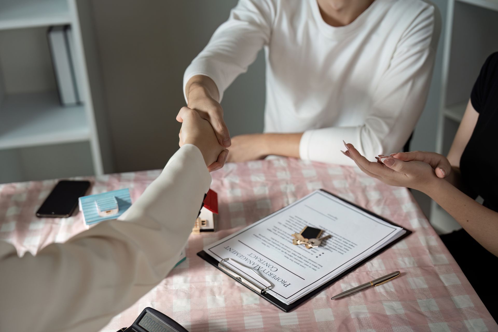 Two people shaking hands over a contract on a table with a model house and a smartphone.