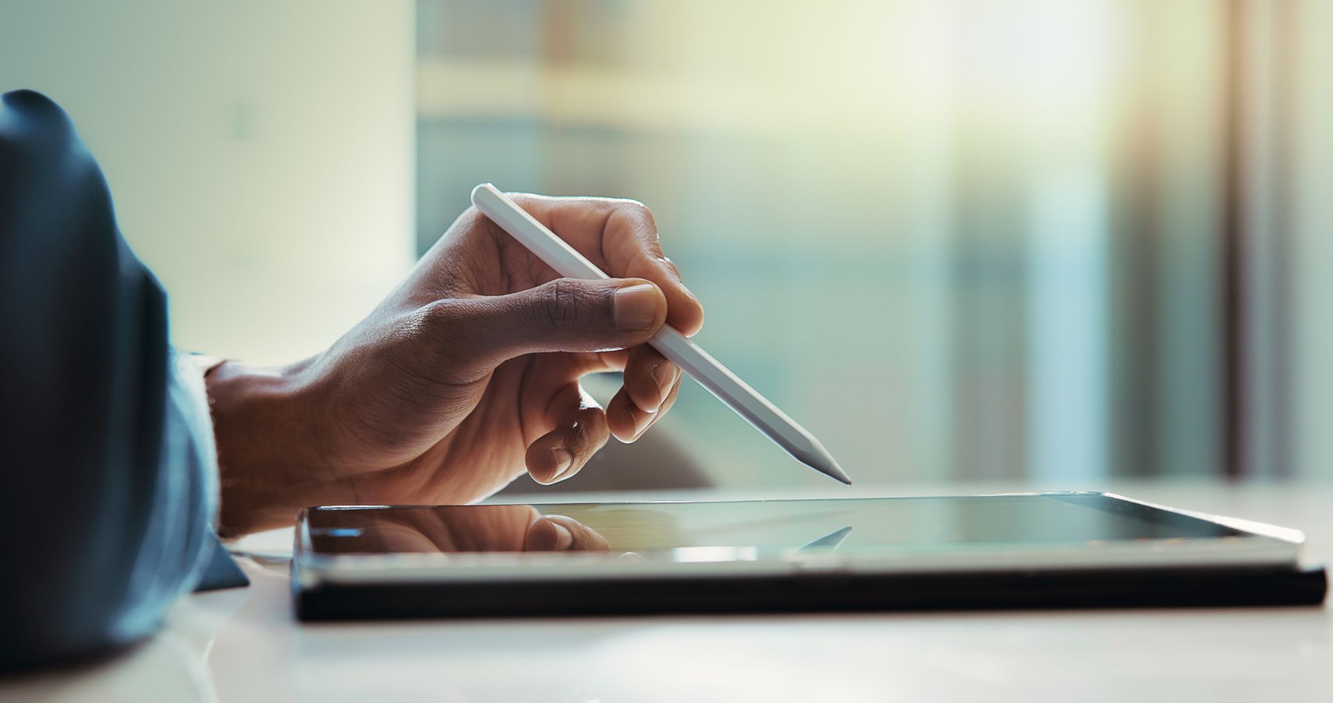 Person's hand using a stylus on a tablet on a table. Sunlight streams in.