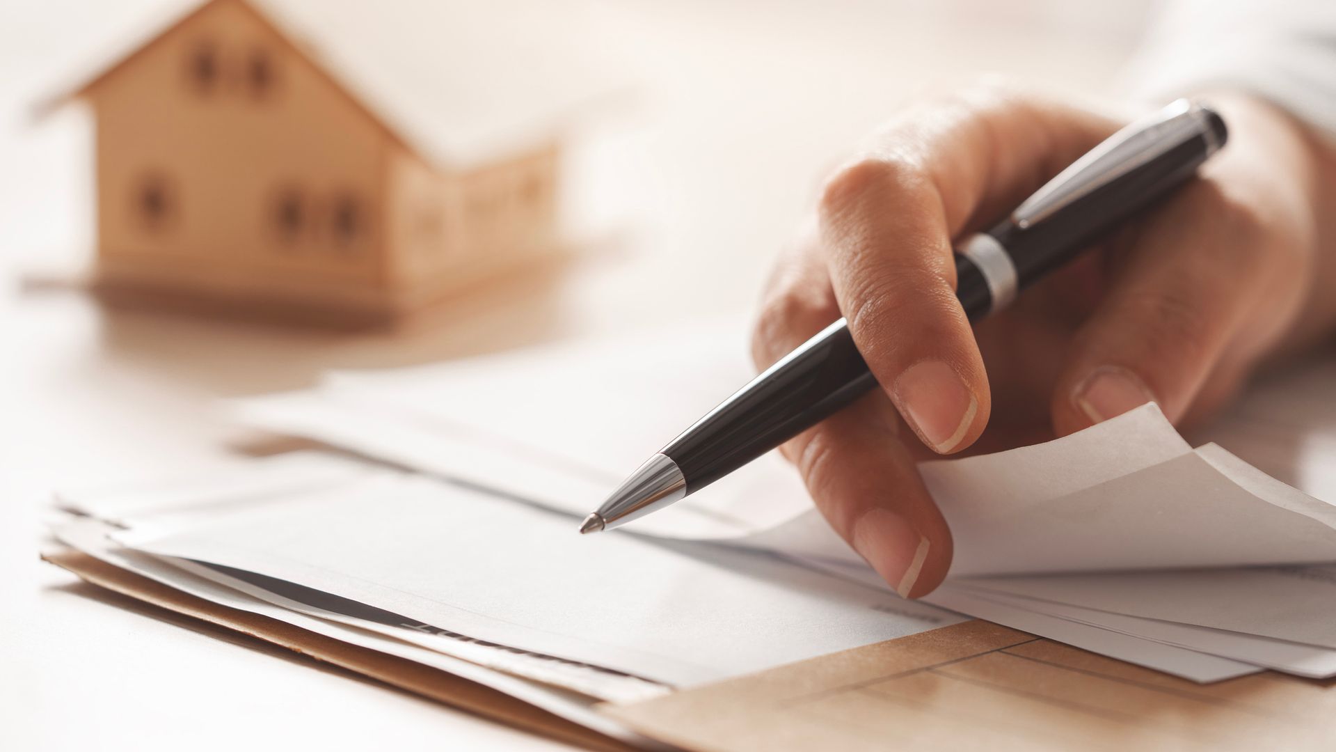 Hand with pen writing on documents near a model house.