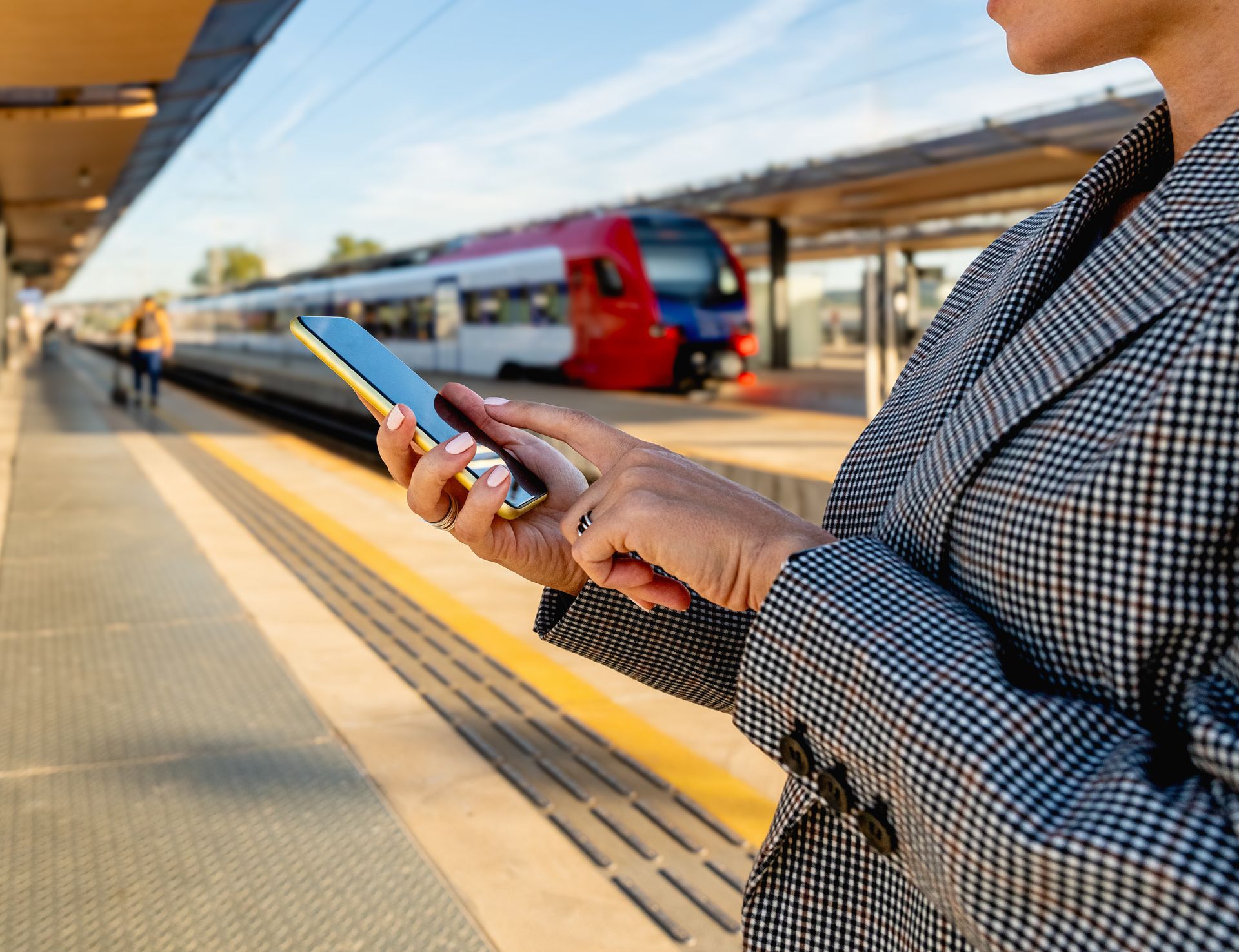Woman in blazer uses phone on train platform, train approaching in background.