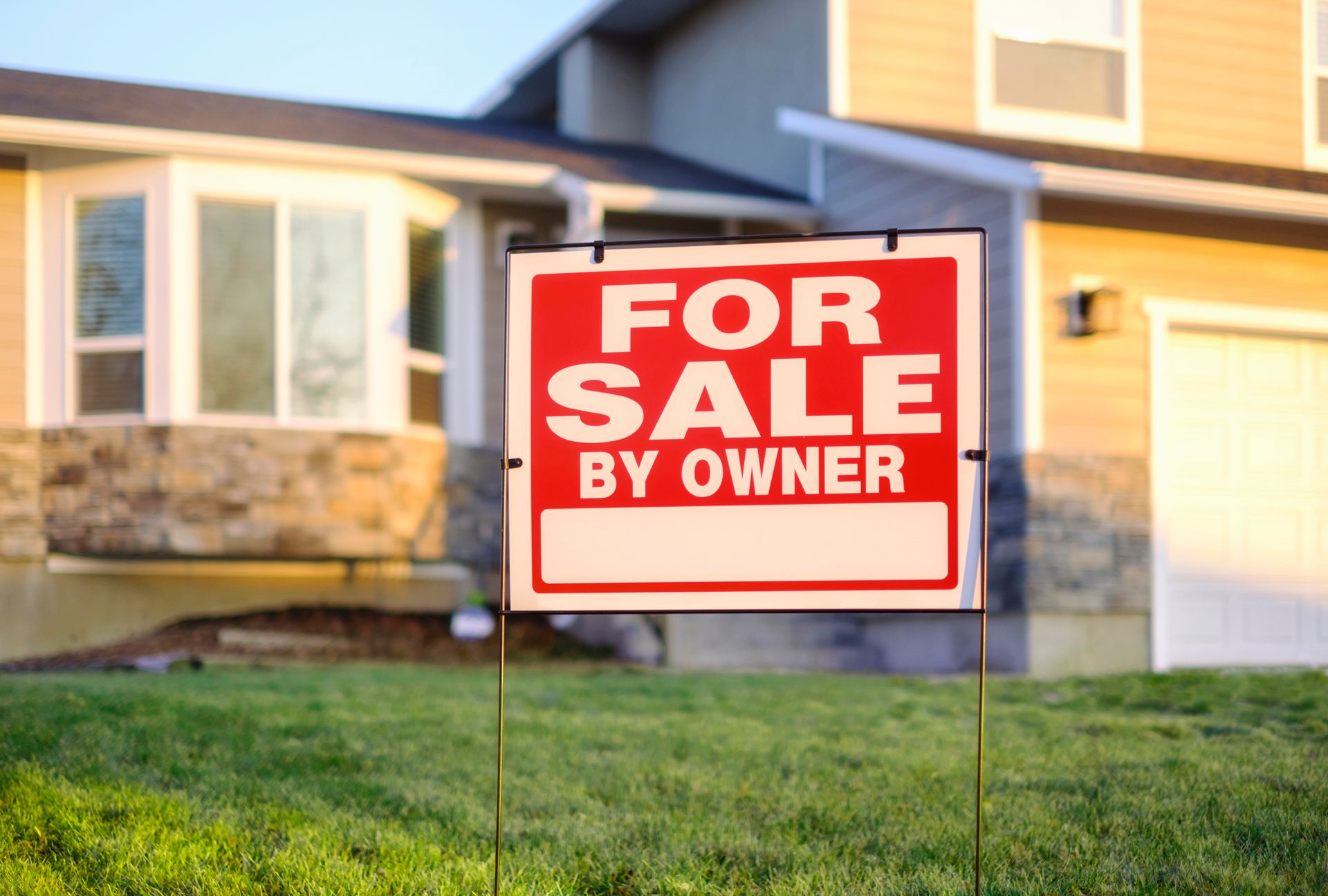 Red For Sale By Owner sign in front of a tan house with a green lawn.