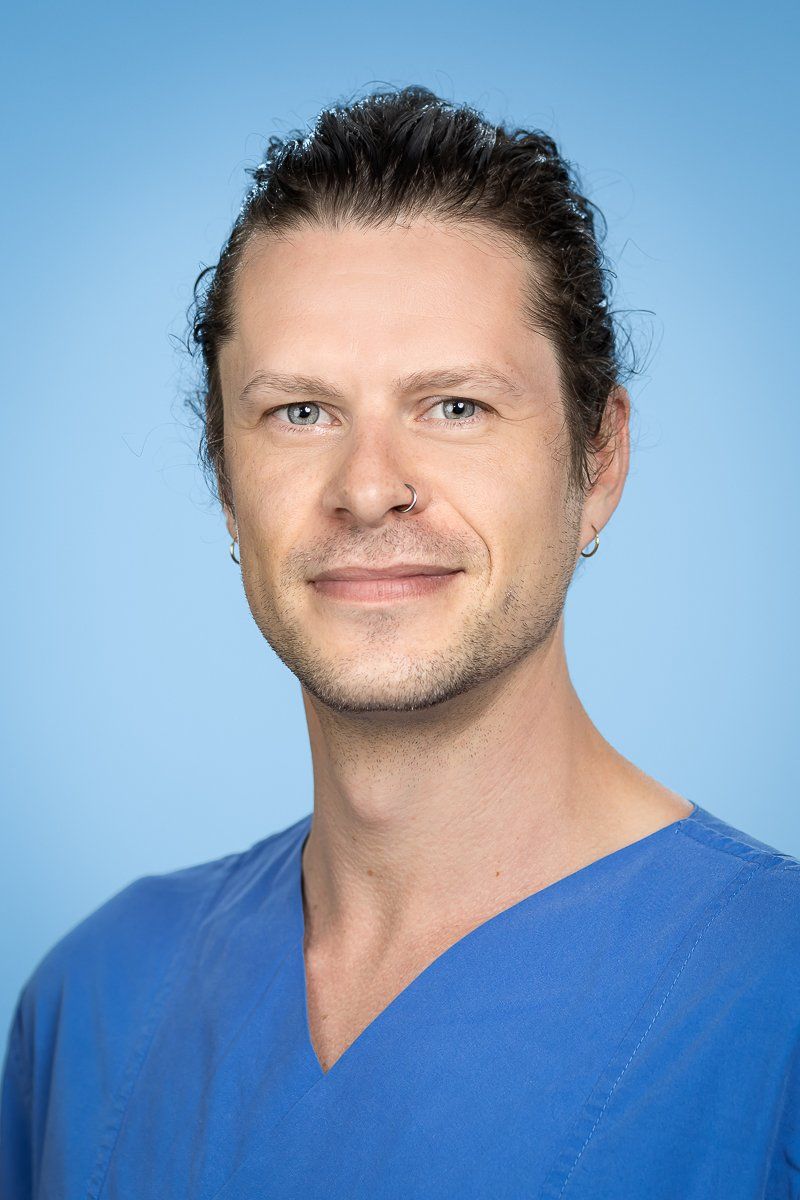 Mann mit zurückgebundenem, dunklem, lockigem Haar, der blaue OP-Kleidung trägt, lächelt in die Kamera.