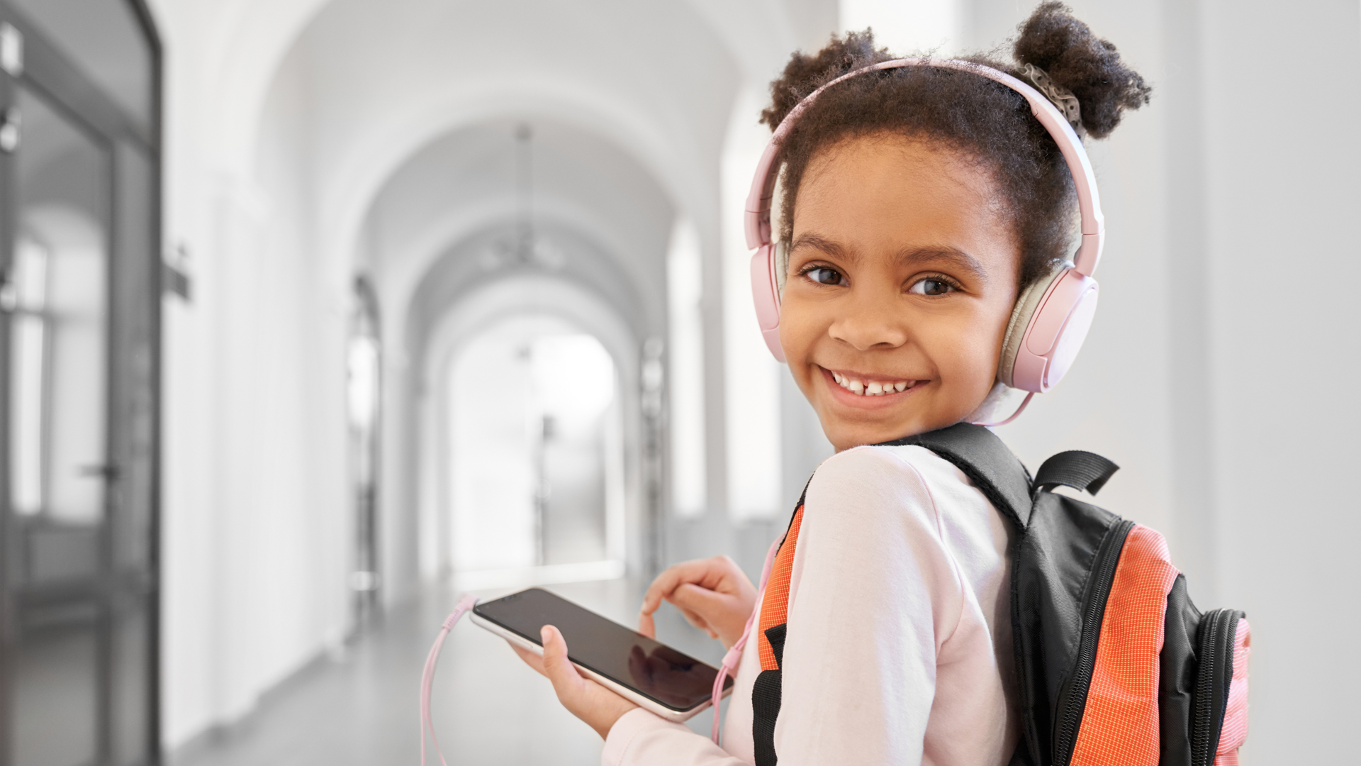 A young girl wearing headphones and a backpack is holding a cell phone in a hallway.