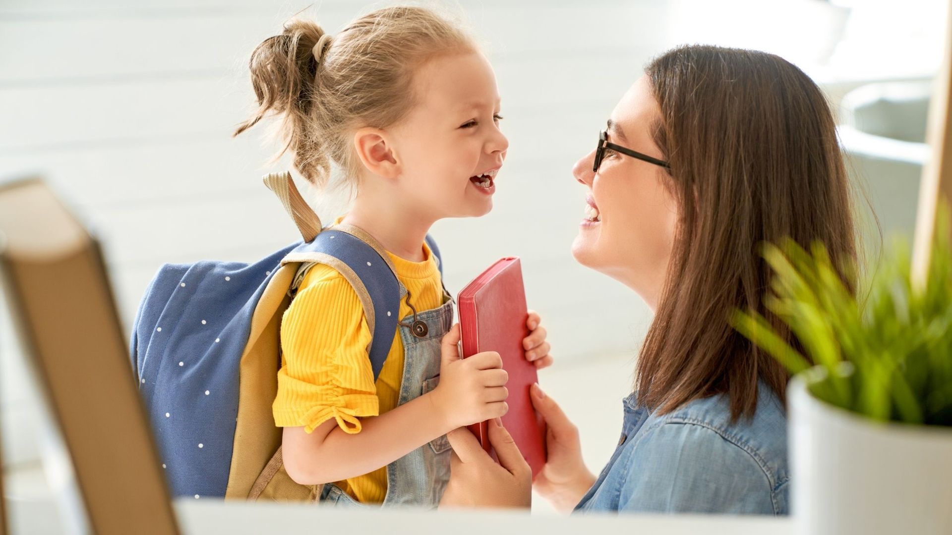 Girl with backpack smiling at woman holding red book; indoor setting.