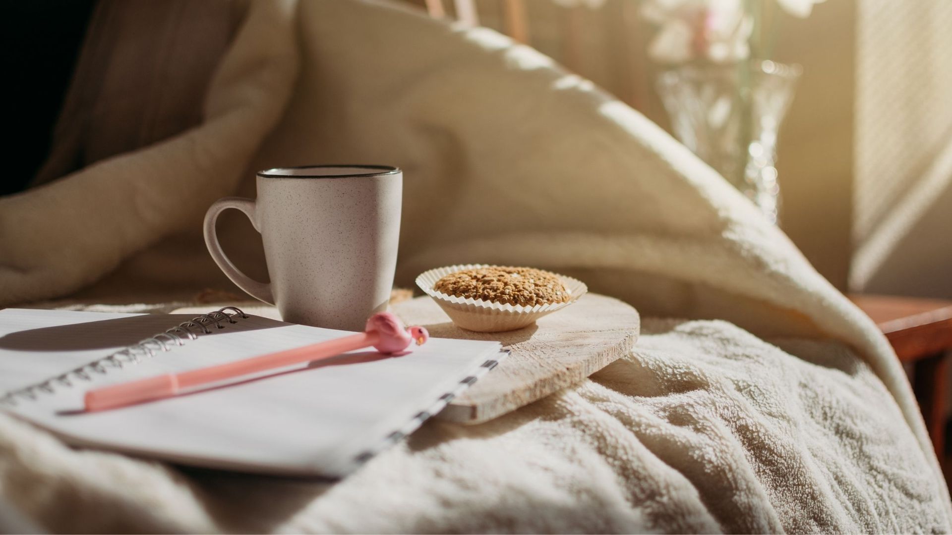 A mug, muffin, pen, and notebook on a bed covered in a blanket, illuminated by sunlight.