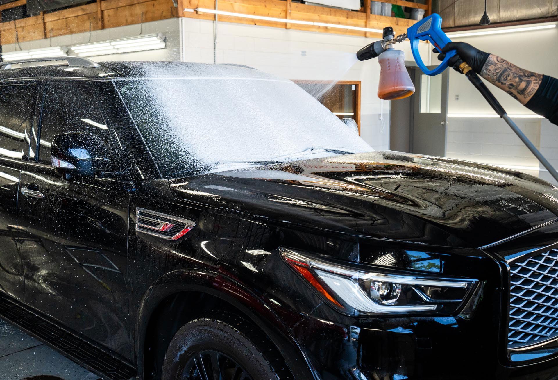 Black SUV being foamed with a detailing sprayer by a person in black gloves.