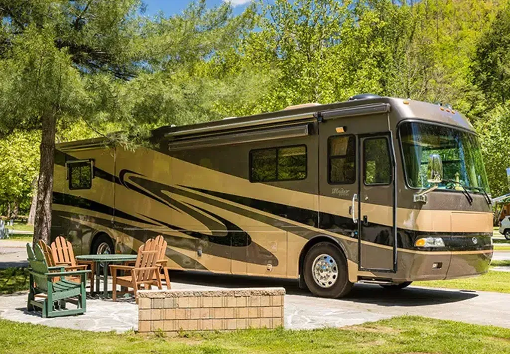 A large rv is parked in a campground with a table and chairs.