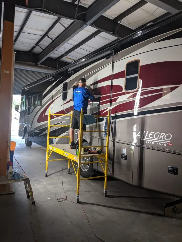 A man is standing on a ladder in front of a rv in a garage.