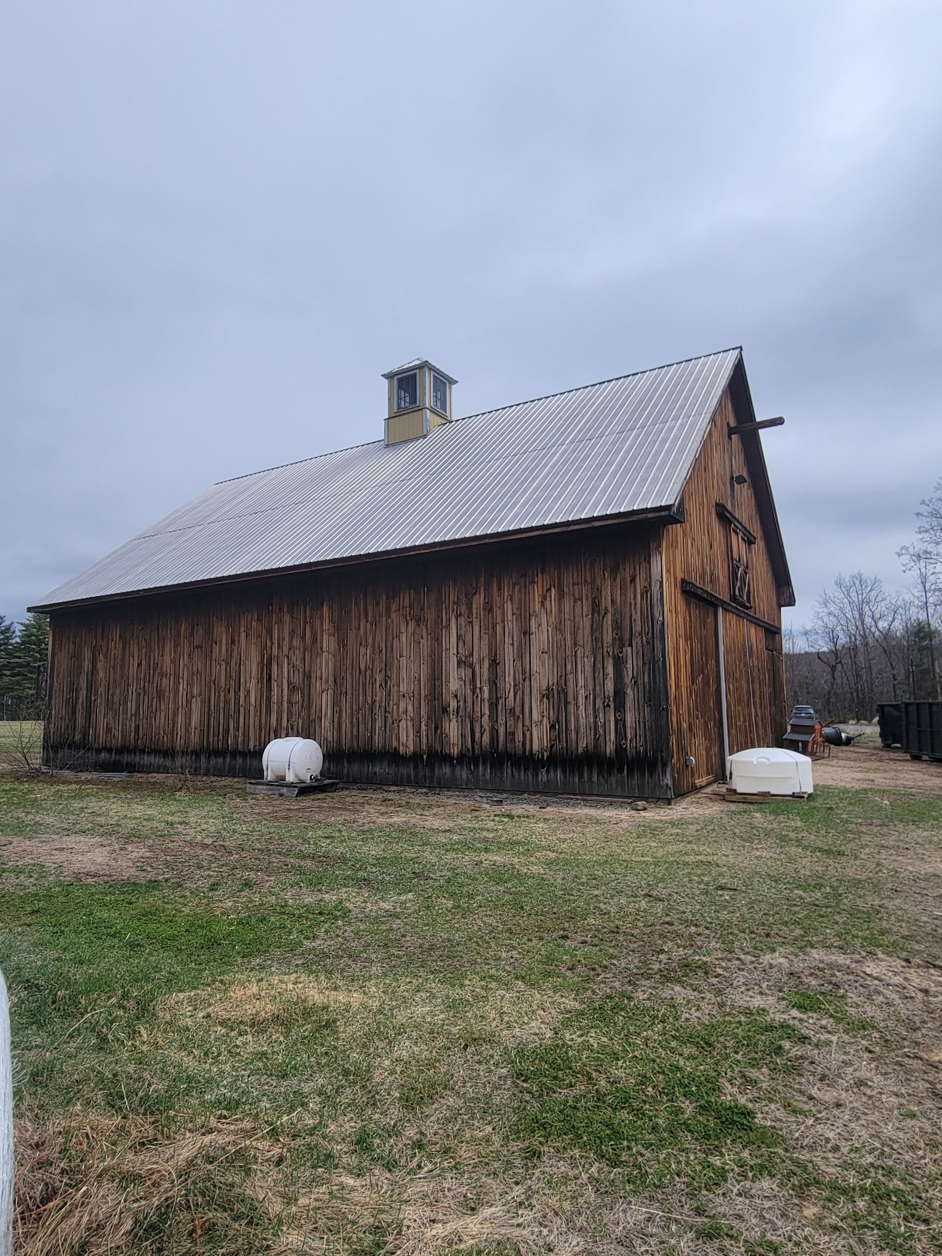A large wooden barn with a metal roof is sitting in the middle of a grassy field.