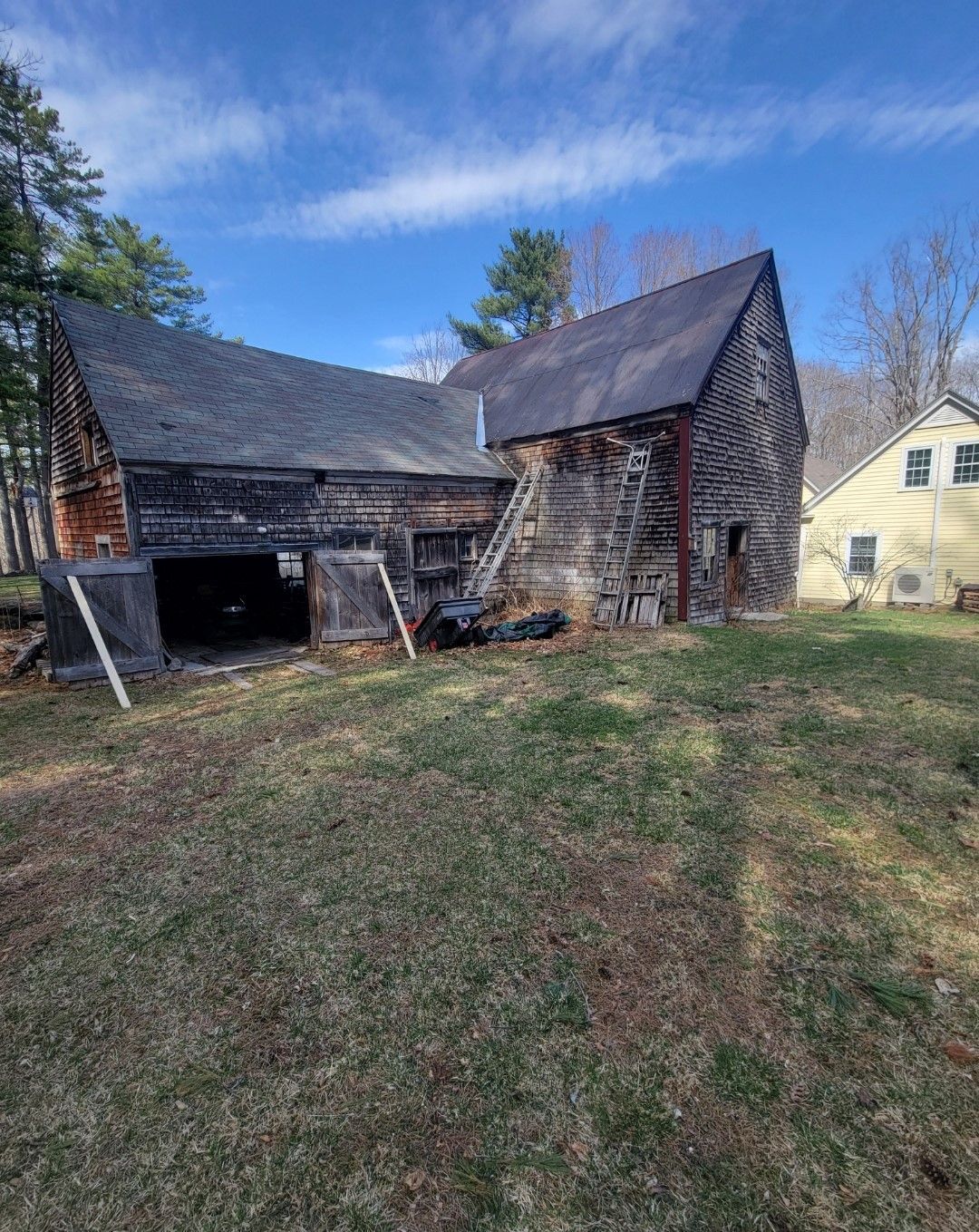 An old barn is sitting in the middle of a grassy field next to a house.