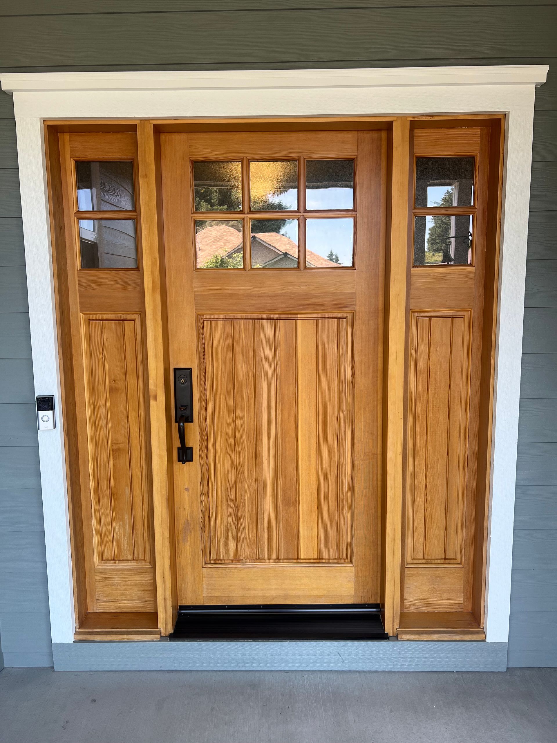 Wooden craftsman style door with sidelights, featuring glass panes, framed by white trim and a black door mat.