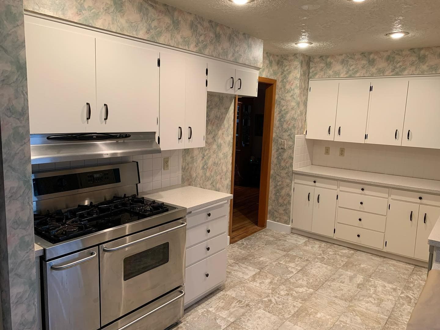 White-cabinet kitchen with a stainless steel stove. Floral wallpaper, tile floor. Doorway.