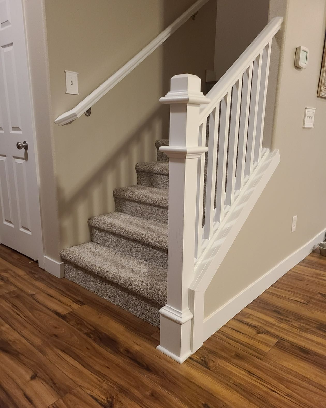 Staircase with carpeted steps, white railing, and wooden flooring. Beige walls with light switches.