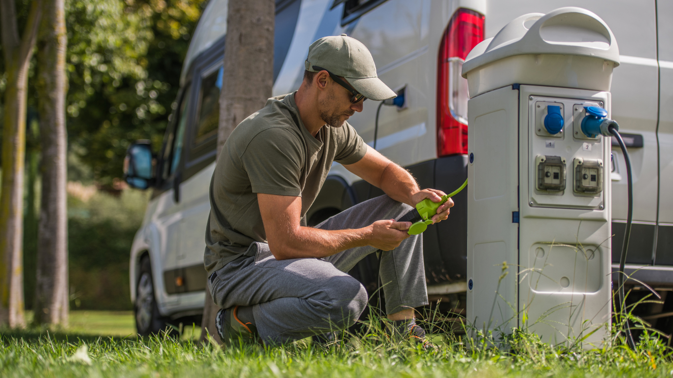 Man in cap kneeling, plugging a green power cord into a campsite electrical box, next to a white campervan on grass.