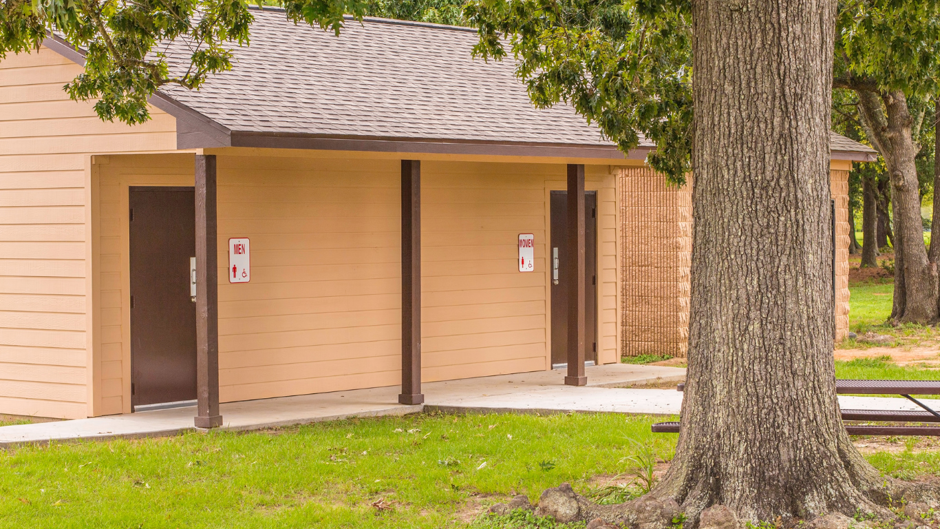 Public restroom building, tan walls, brown doors, under a tan roof, in a grassy area. A tree trunk is in the foreground.