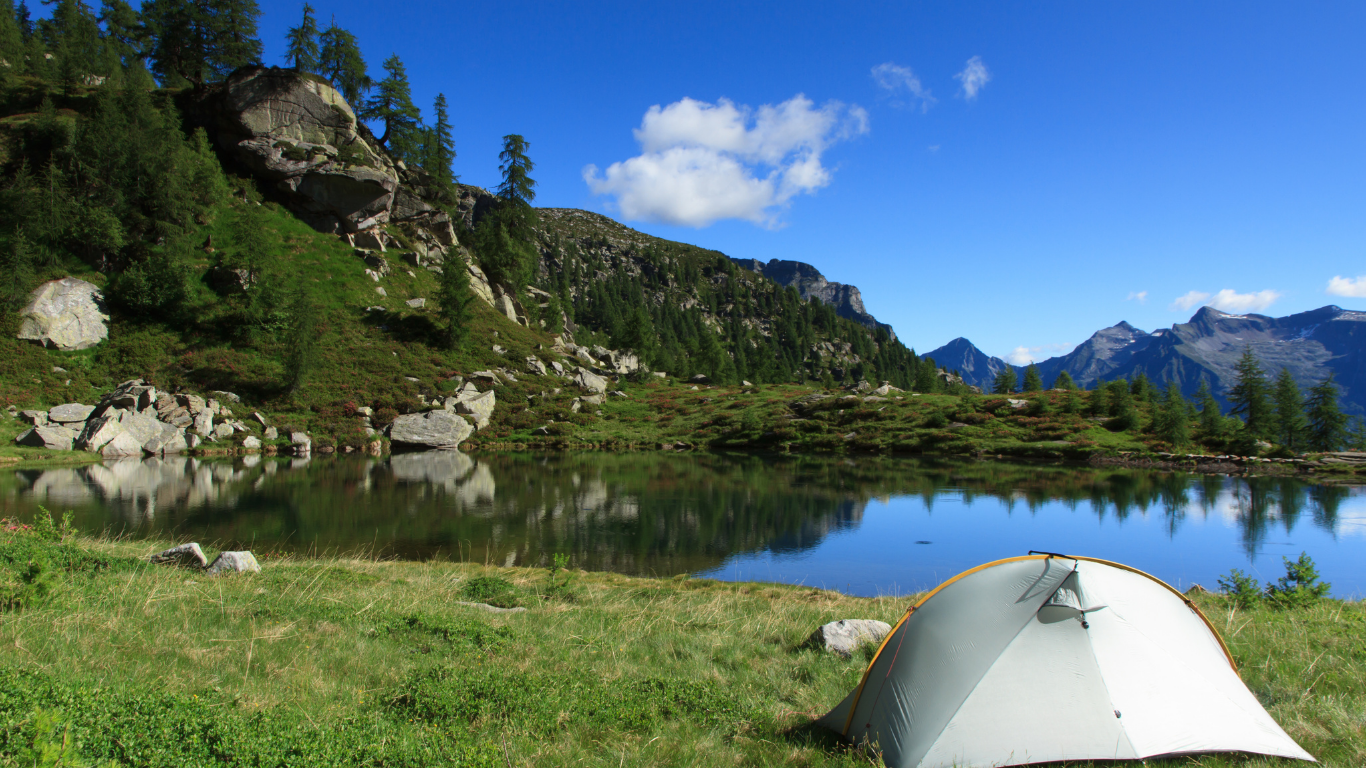 A small, white tent pitched beside a calm lake with mountains and blue sky in the background.