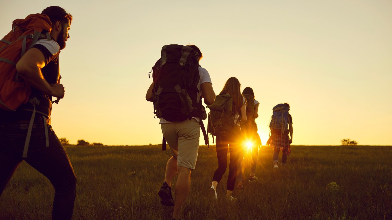 Group of hikers with backpacks walking towards the sunset in a grassy field.