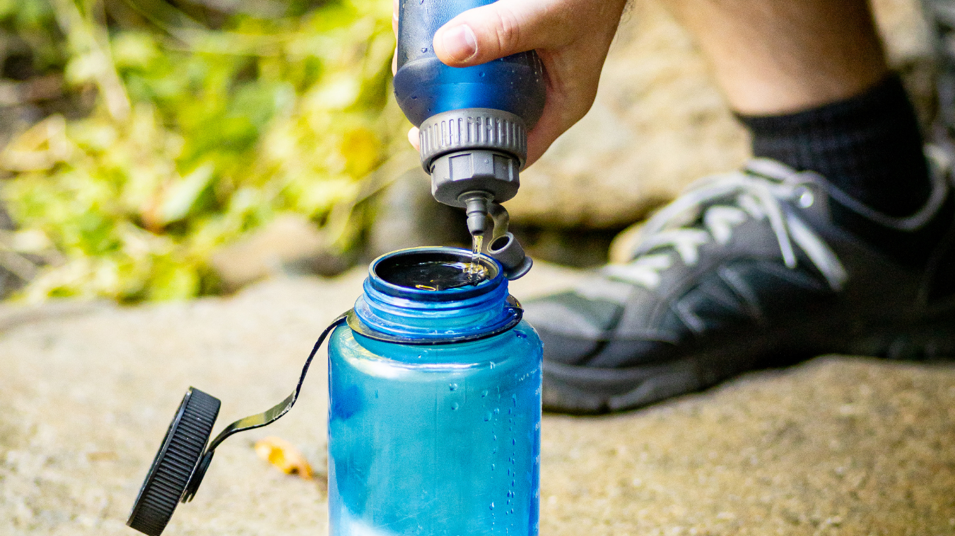 Person pouring liquid from a blue bottle into a larger blue water bottle outdoors.