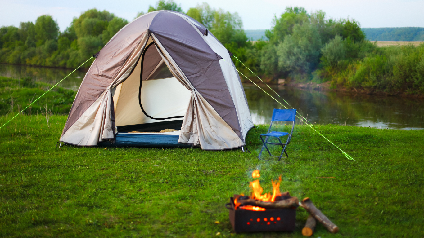 Camping scene: tent set up near a river with a small fire, blue folding chair, and green grass.
