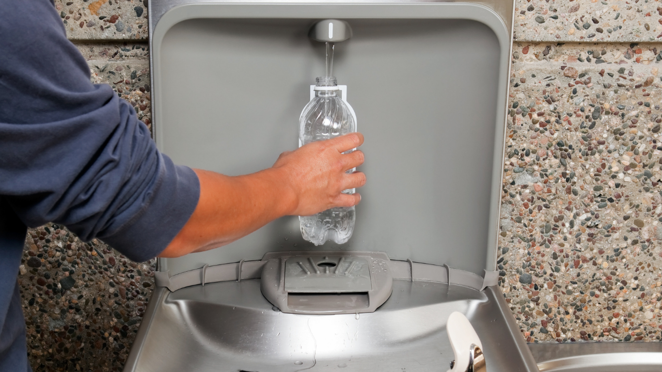 Person filling a plastic water bottle from a water fountain, set against a grey and speckled wall.