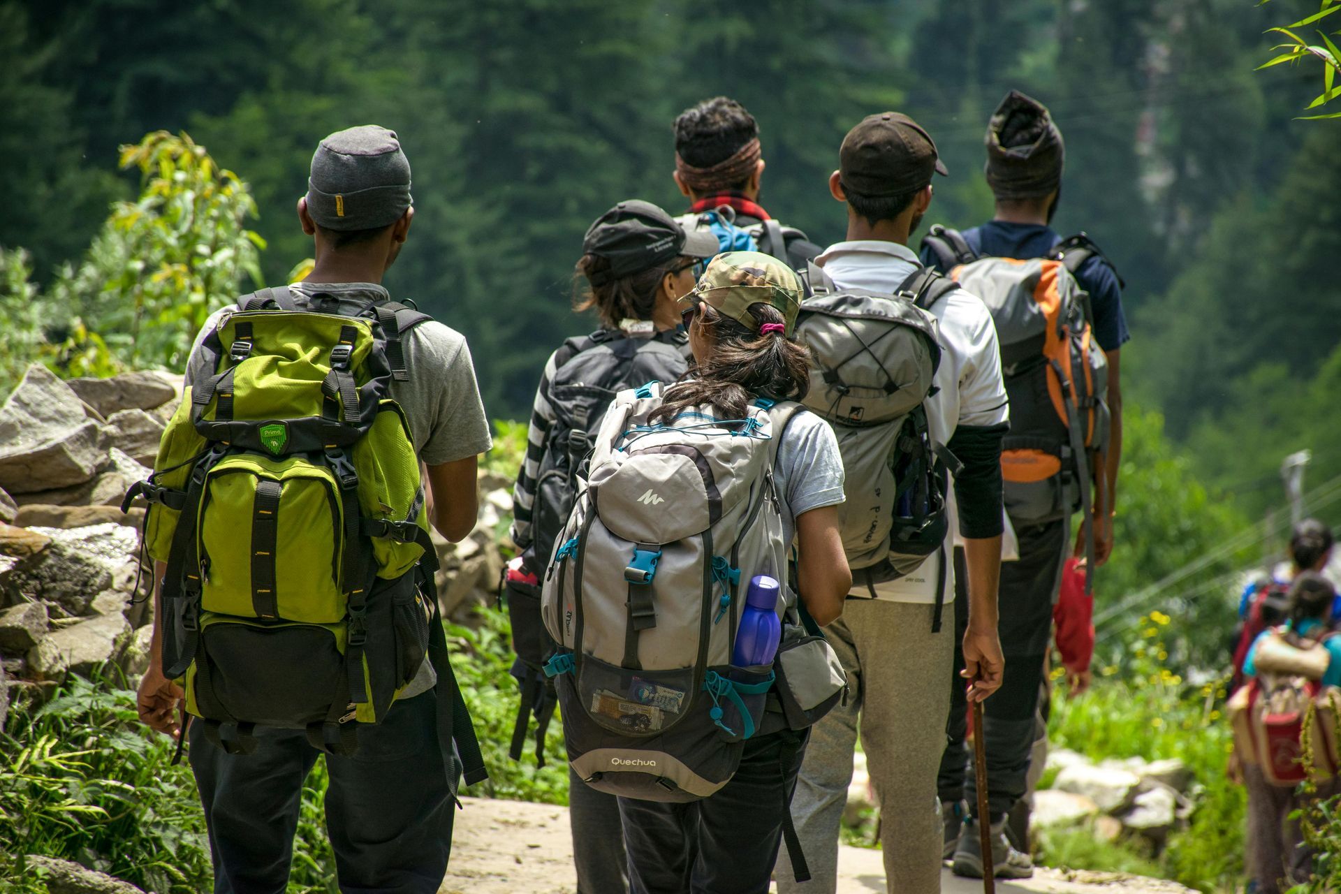 Group of hikers with backpacks trekking on a trail in a green, mountainous area. They are walking away from the camera.