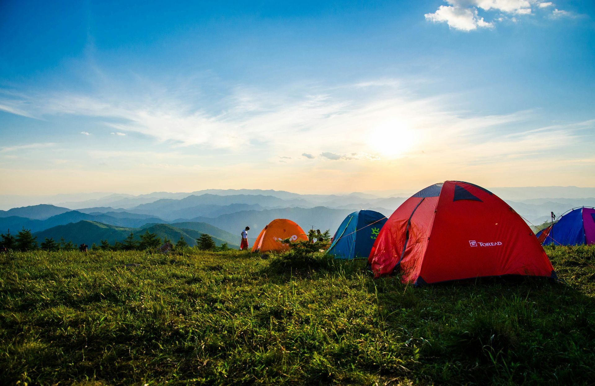 Camping tents on a grassy mountaintop at sunrise. The sky is blue with the sun's light shining.