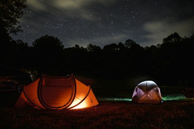 Two illuminated tents in a grassy field at night under a starry sky; trees in the background.