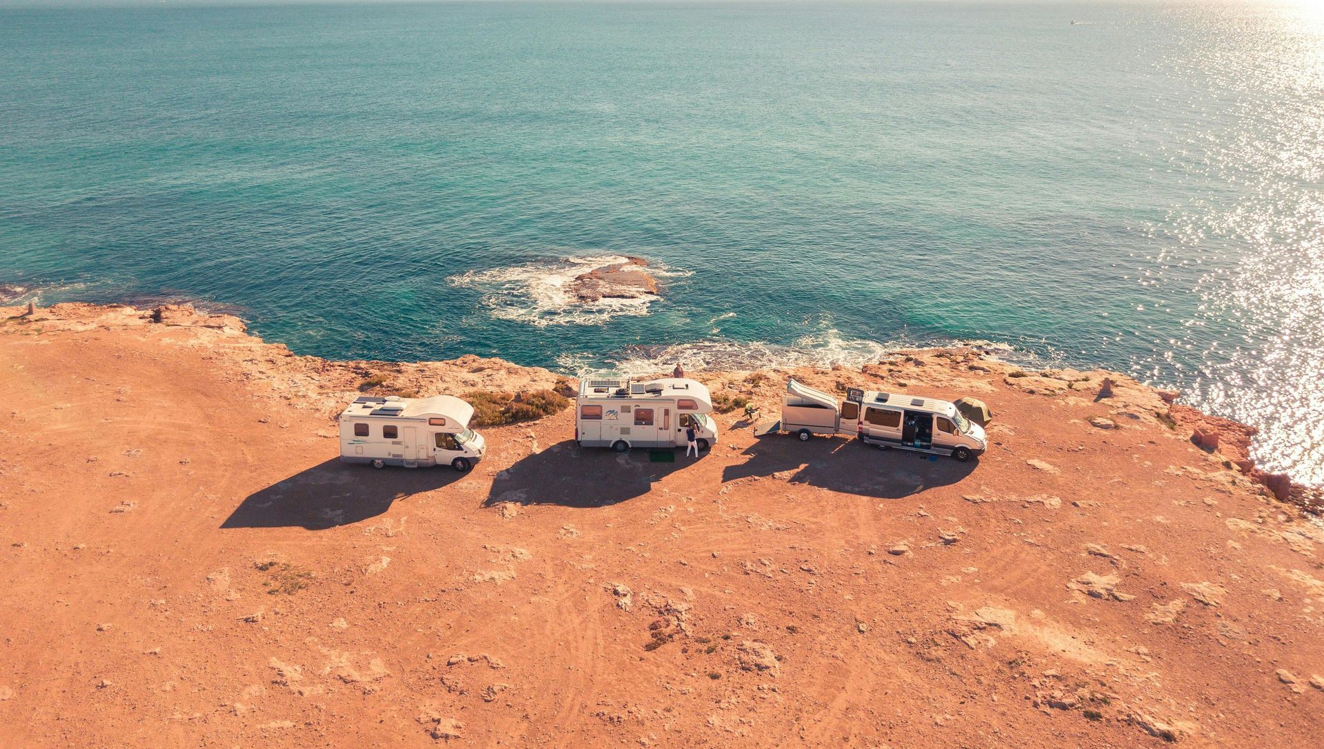 Three RVs parked on a red-brown cliff overlooking a turquoise ocean. Sunlight glints on the water.