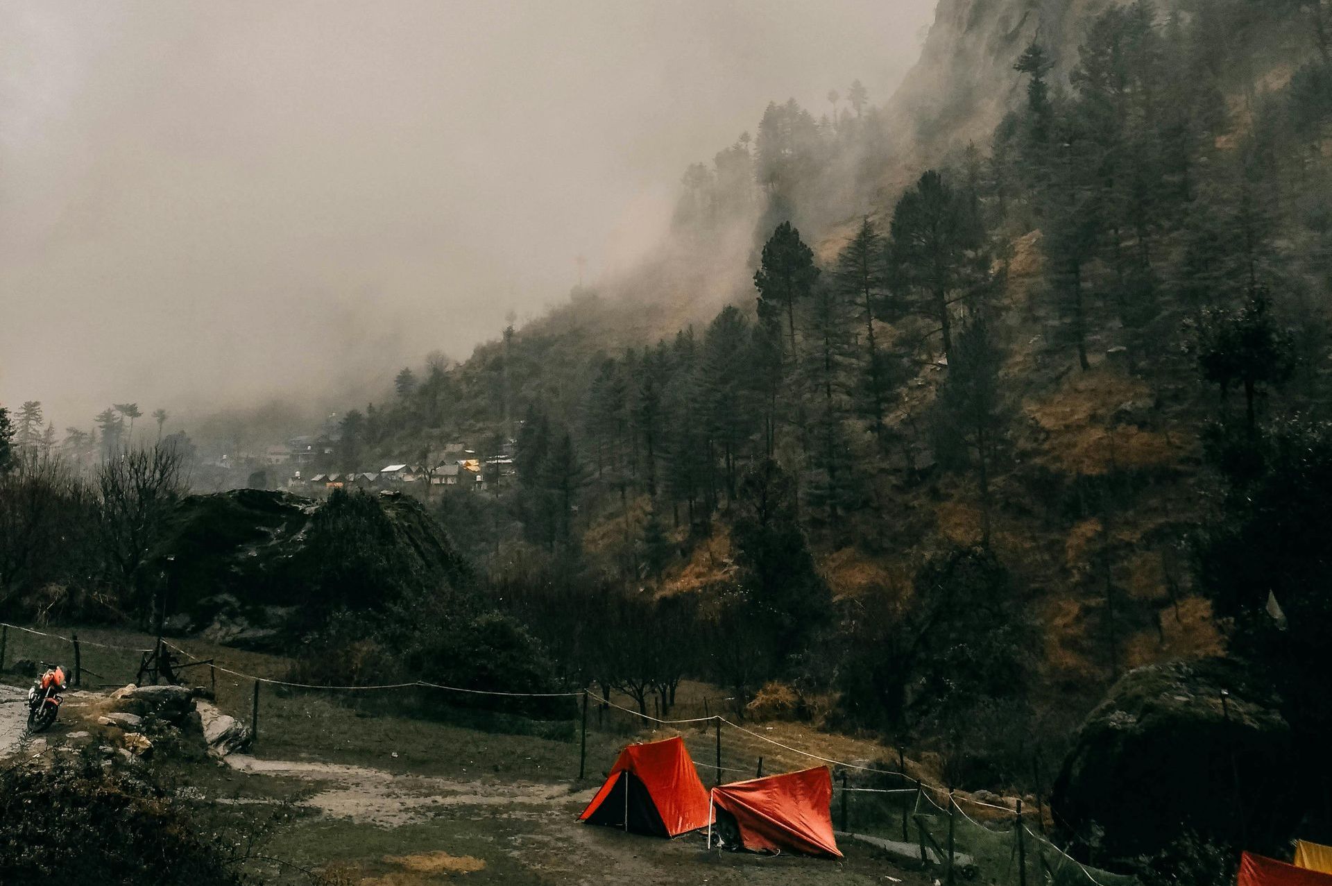 Camping tents in a grassy field at the foot of a misty mountain. Dark trees dot the hillside under a cloudy sky.
