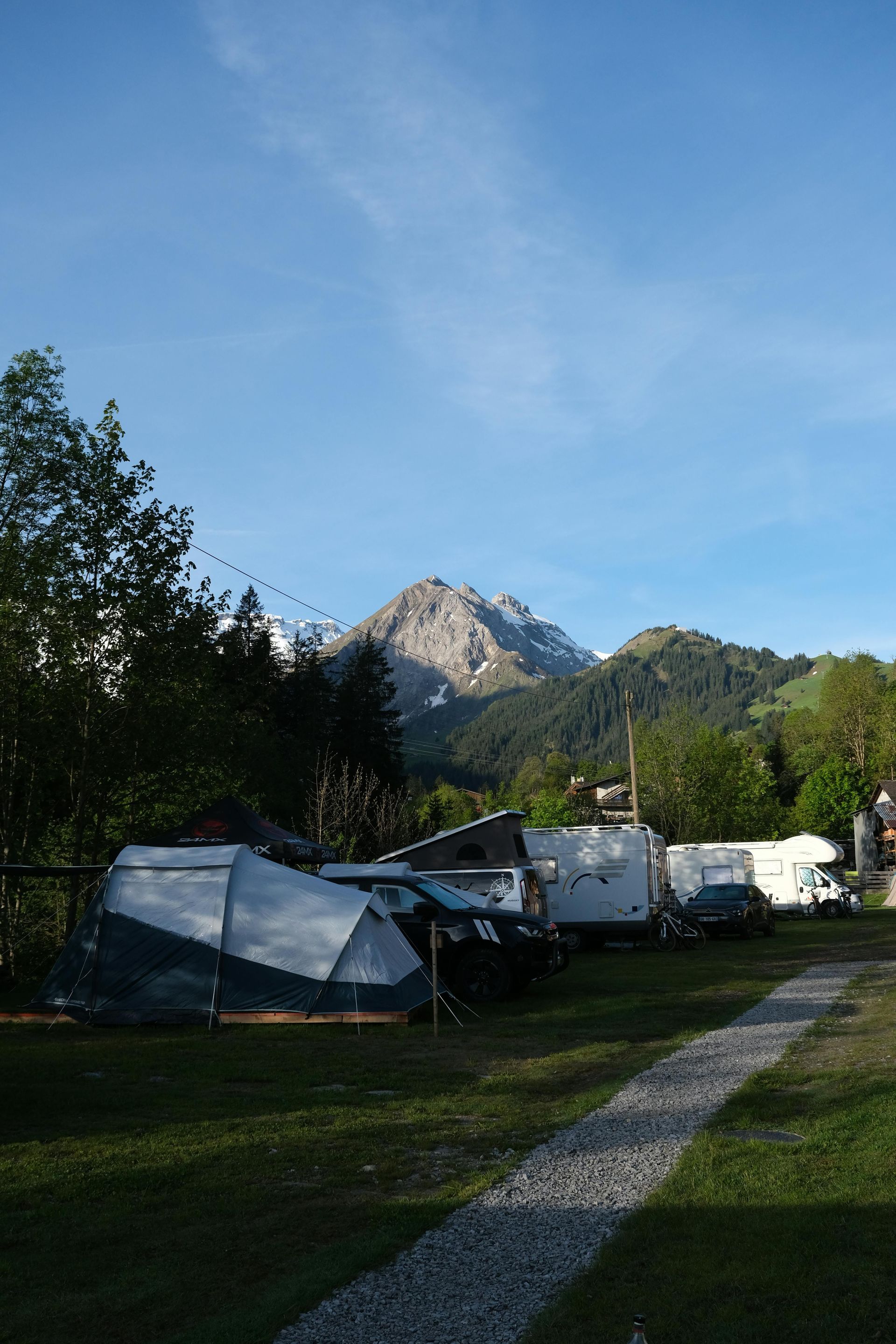 Camping scene with tents and RVs in front of a mountain under a blue sky. A gravel path leads toward the campsite.