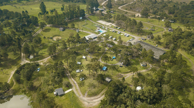 Aerial view of a campsite nestled amidst green trees and fields, featuring tents, buildings, and a dirt road.
