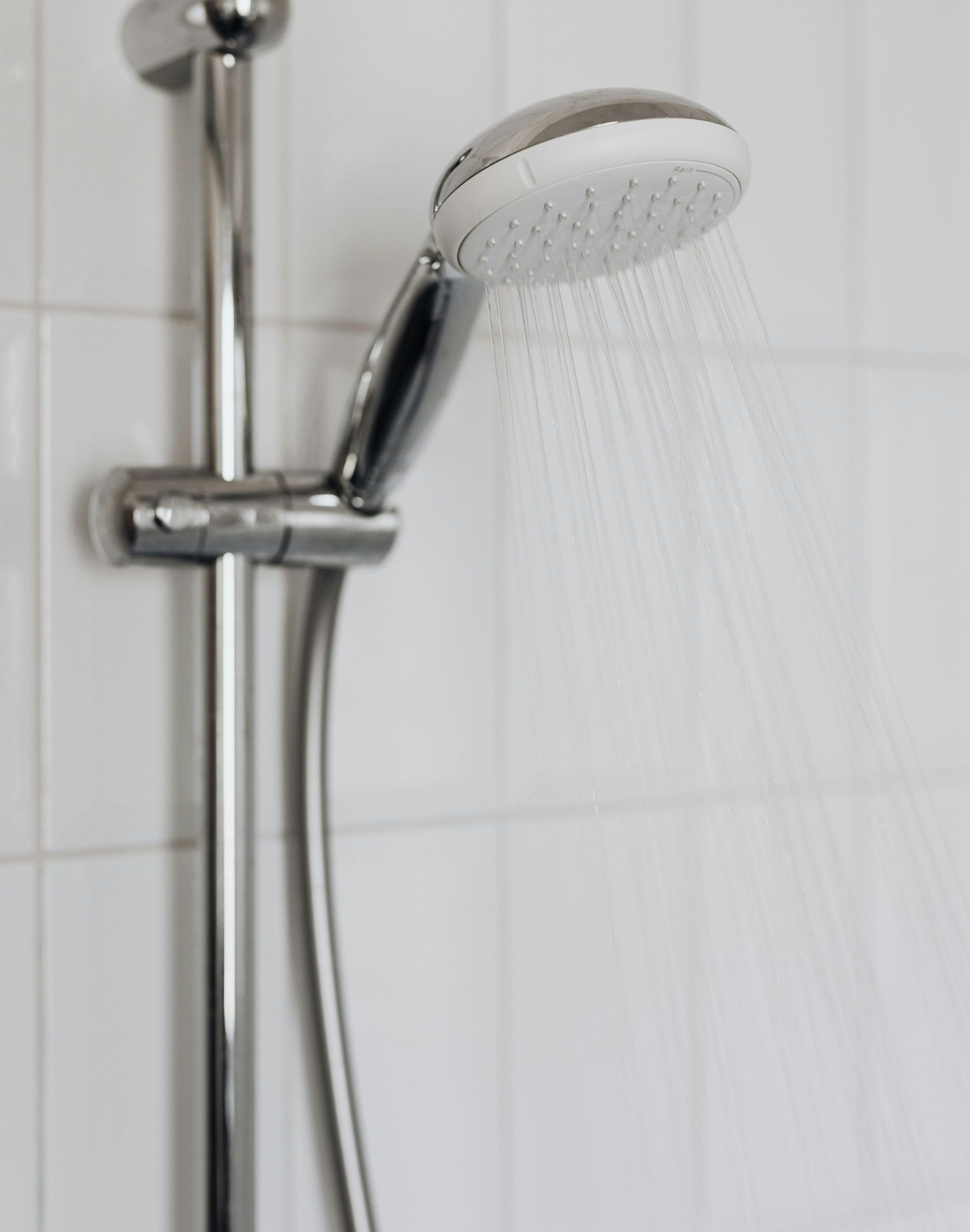 Close-up of a chrome showerhead with water flowing, mounted on a vertical bar against white tiled wall.