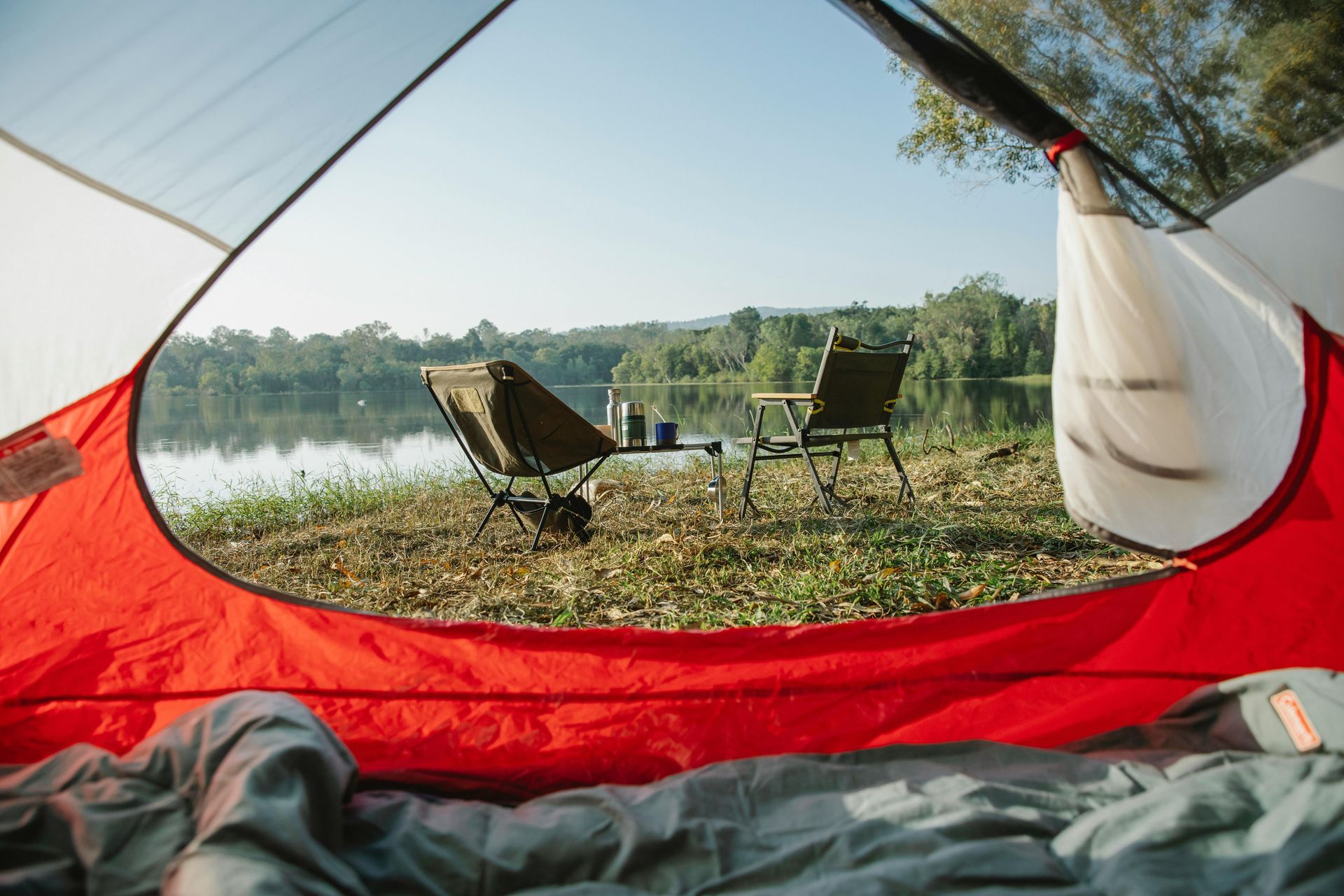 View from inside a red tent overlooking a lake with two camping chairs and a small table.