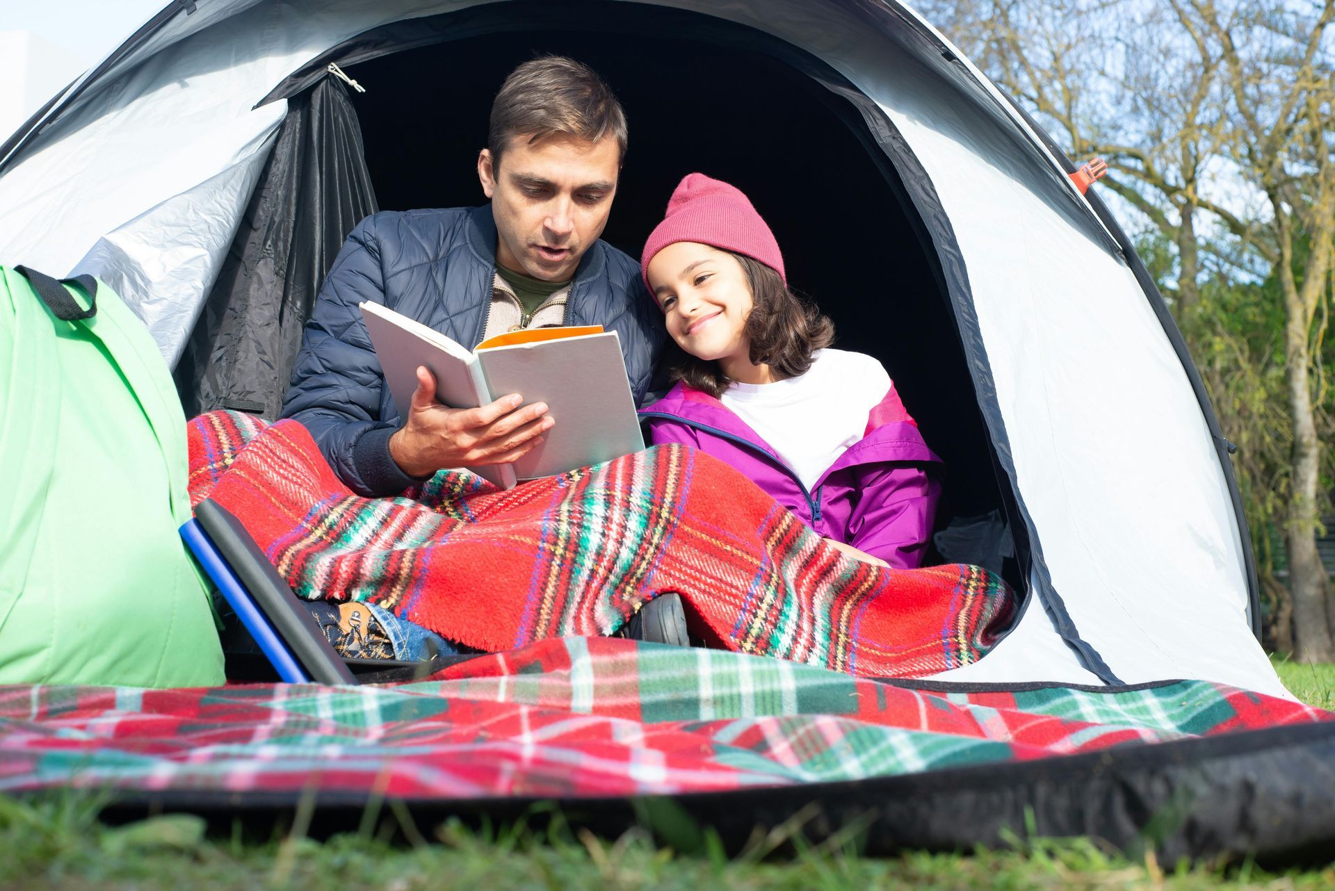 A man and child sit inside a tent, reading a book. The child, smiling, wears a pink hat and purple coat.