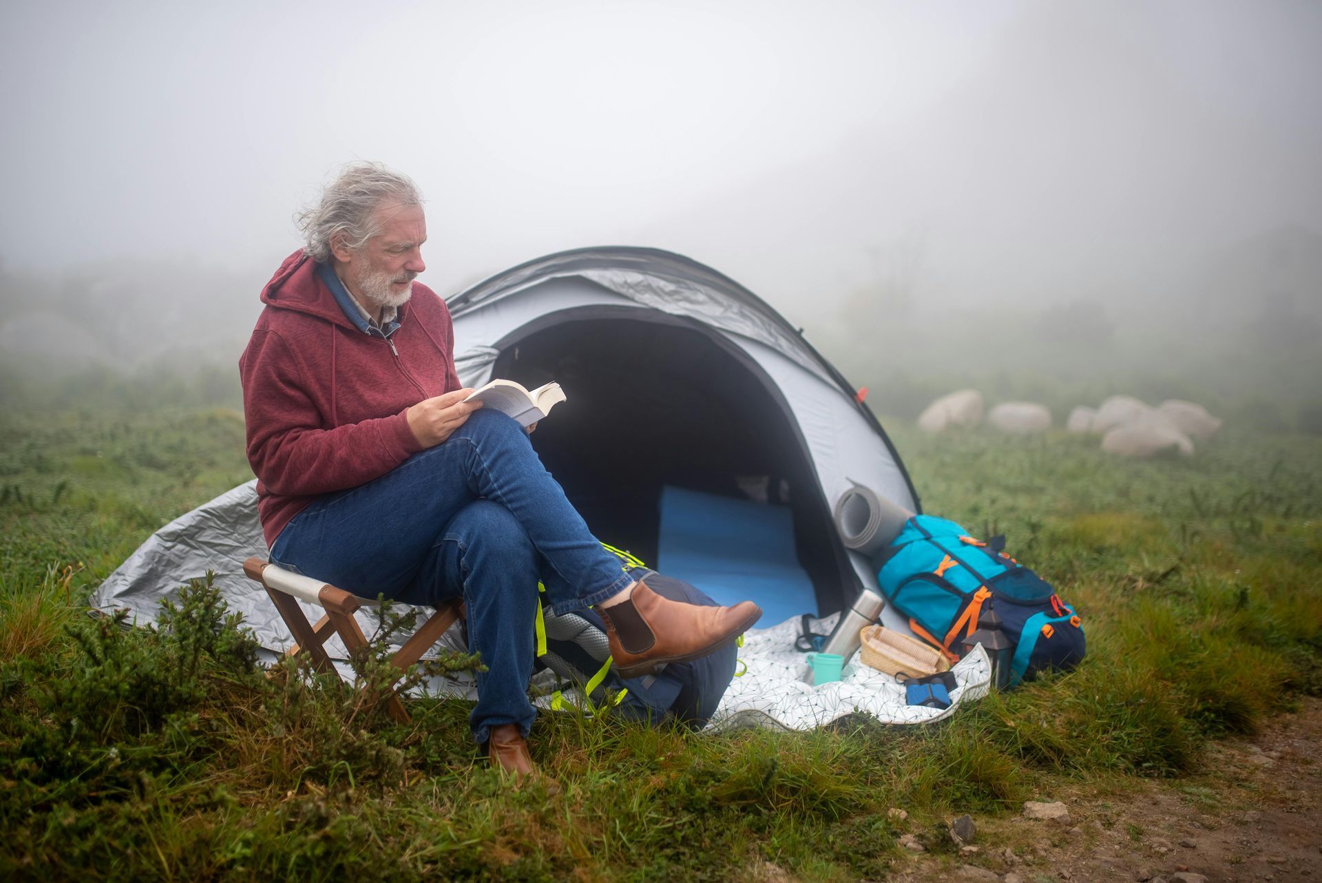 A man with a beard reads outside a tent in a foggy field. He wears a red jacket, blue jeans, and brown boots.