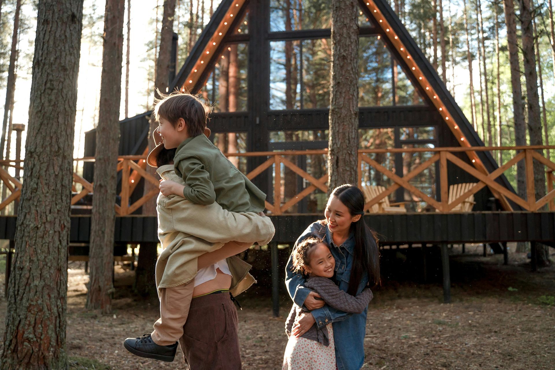 Family in a forest setting. A man holds a child up; a woman hugs another child in front of a modern cabin.