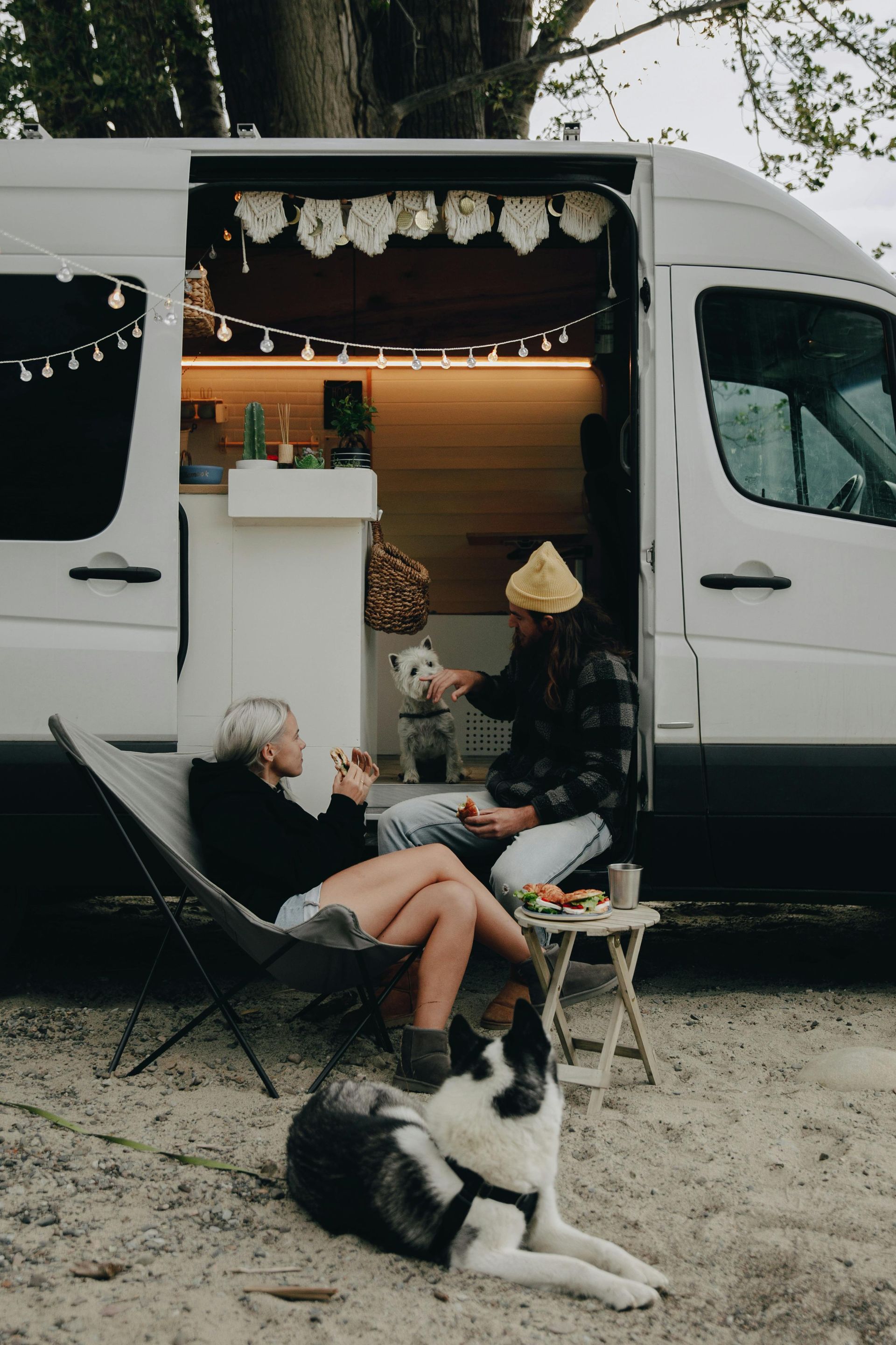 A couple and a dog relax outside a camper van. The woman is sitting in a chair, and the man feeds a dog.