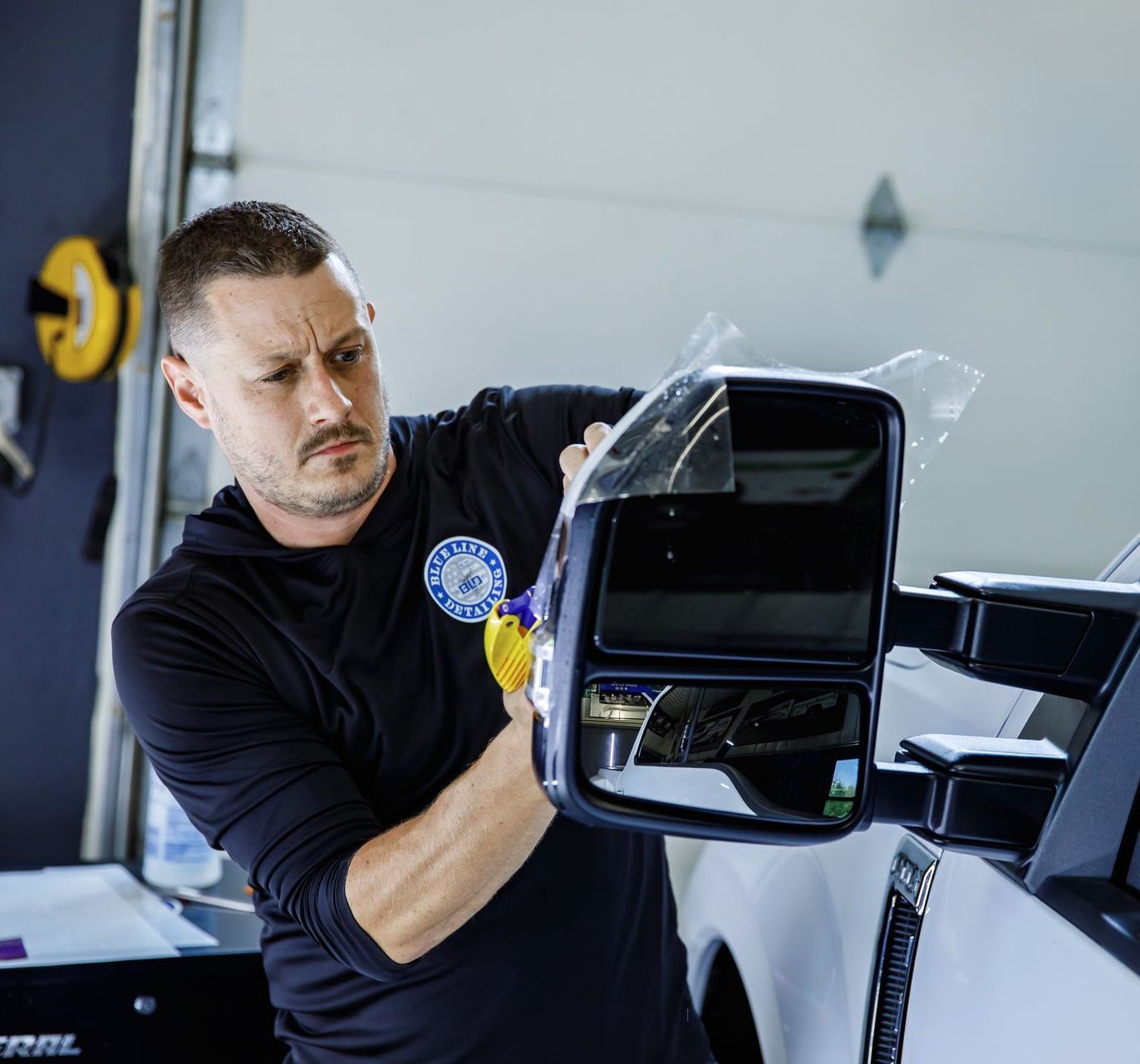 Man applying tint to a truck mirror in a garage. He wears a black shirt, looks focused.
