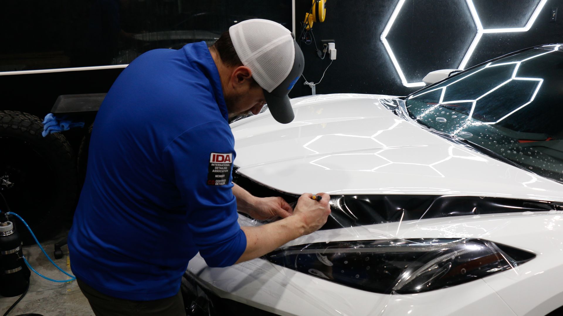 Man in blue shirt applies film to a white car hood in a shop with hexagonal lights.