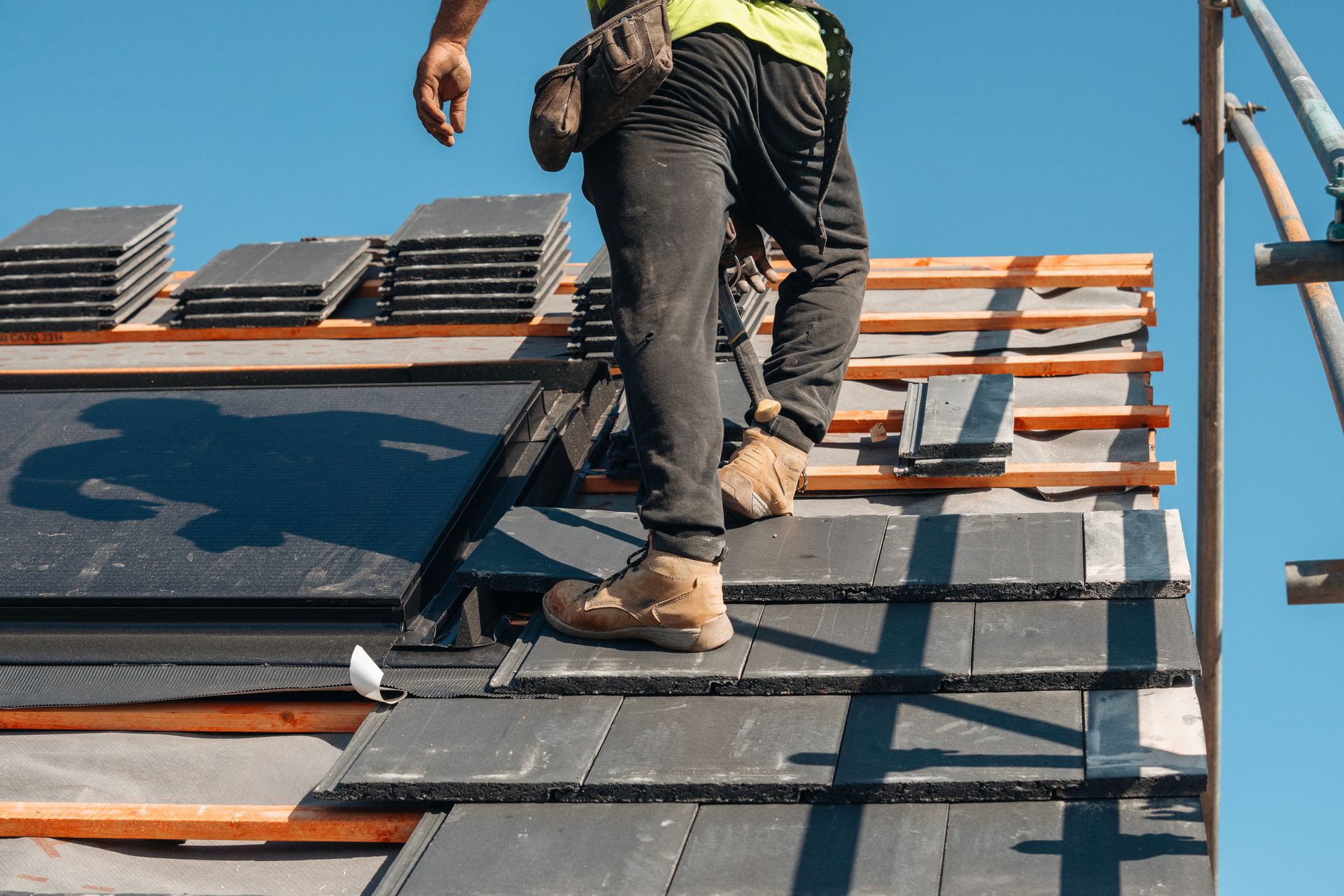 Roofer laying dark gray tiles on a partially constructed roof under a blue sky. Roofer laying dark gray tiles on a partially constructed roof under a blue sky.