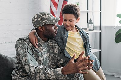 Soldier in camo uniform talking with child; both smiling, indoors, American flag in background.