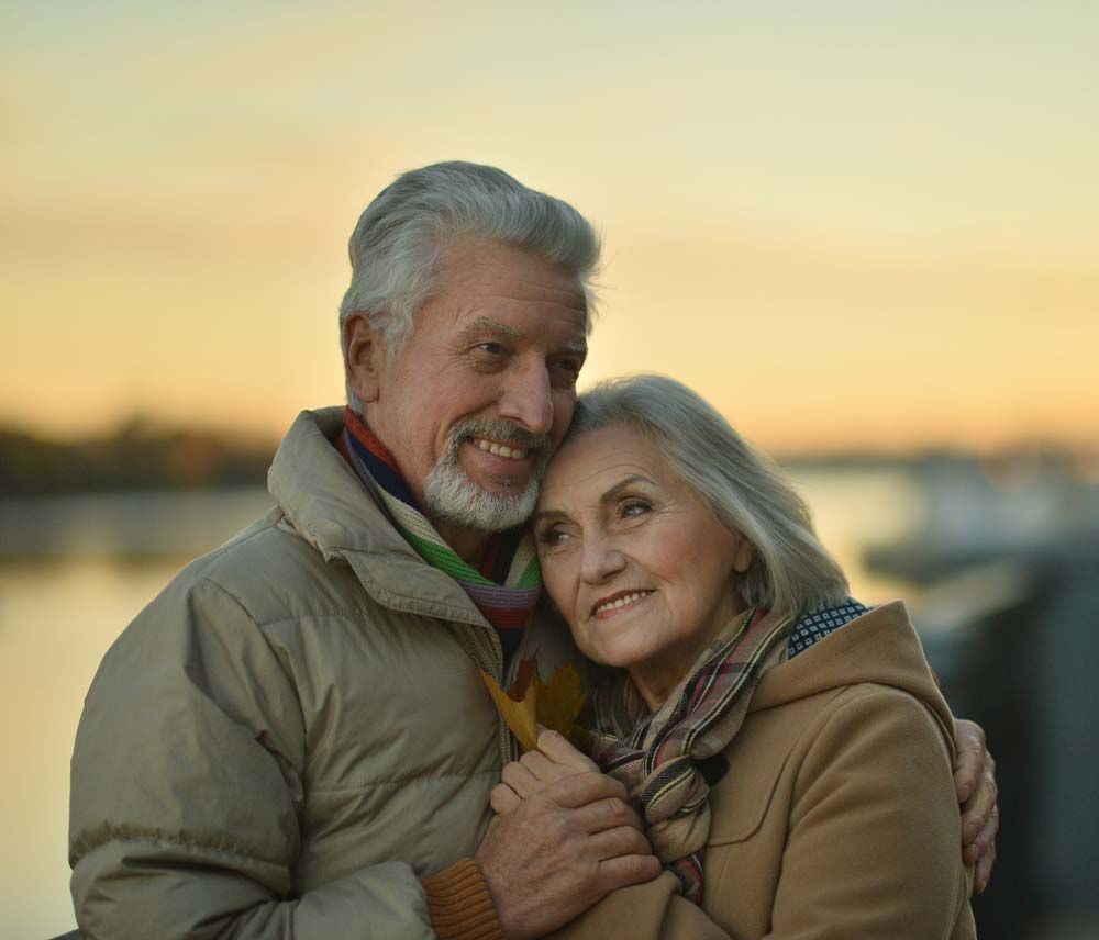 An elderly couple is hugging each other in front of a body of water.