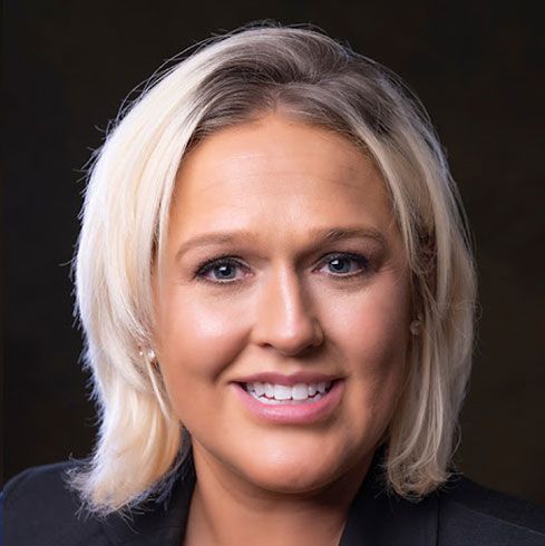 Woman with blonde hair smiles, wearing a black blazer, looking directly at the camera.