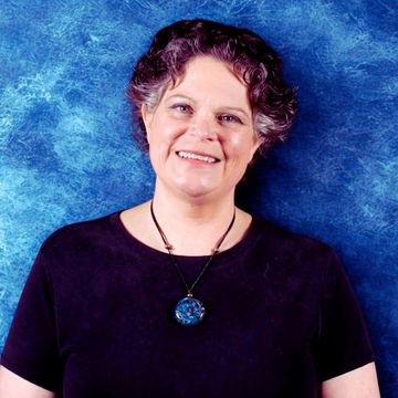 A photo of Ginny Hicks, Licensed Massage Therapist, in a black shirt smiling against a blue background
