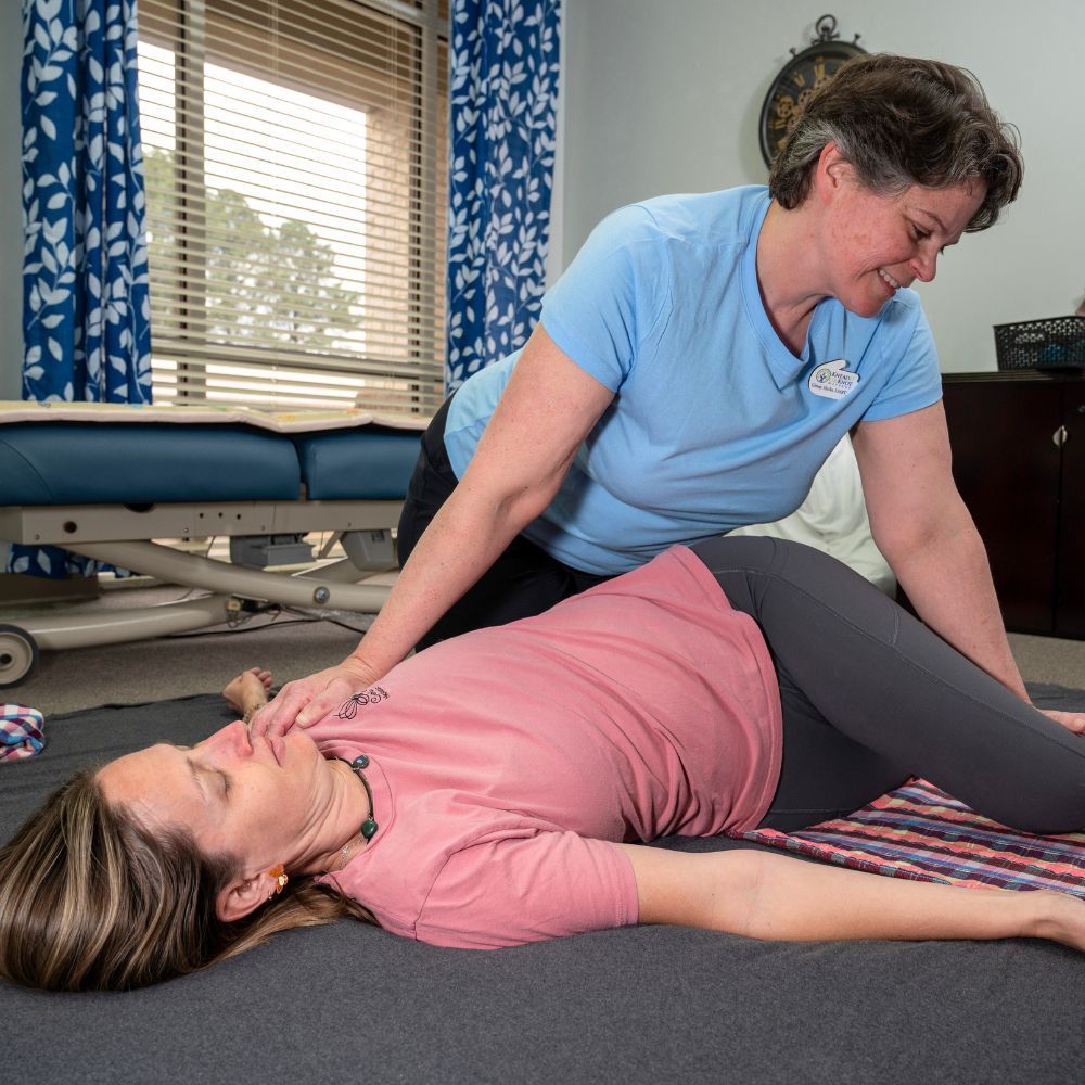 A woman (Ginny Hicks, LMBT) is stretching a client receiving a Thai yoga massage on a mat on the floor.