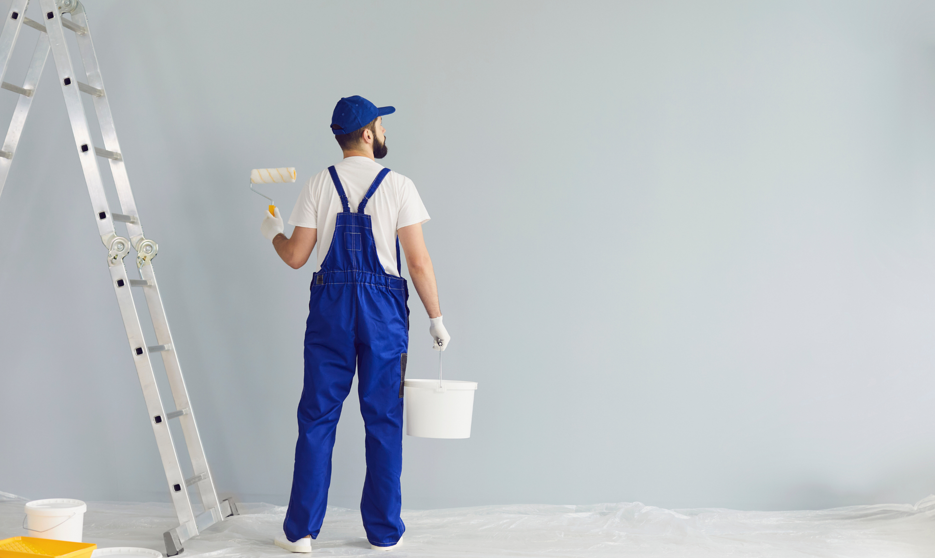 Painter in blue overalls with a paint roller, standing near a ladder, painting a wall. Painter in blue overalls with a paint roller, standing near a ladder, painting a wall.