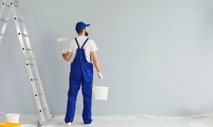 Painter in blue overalls with a paint roller, standing near a ladder, painting a wall.