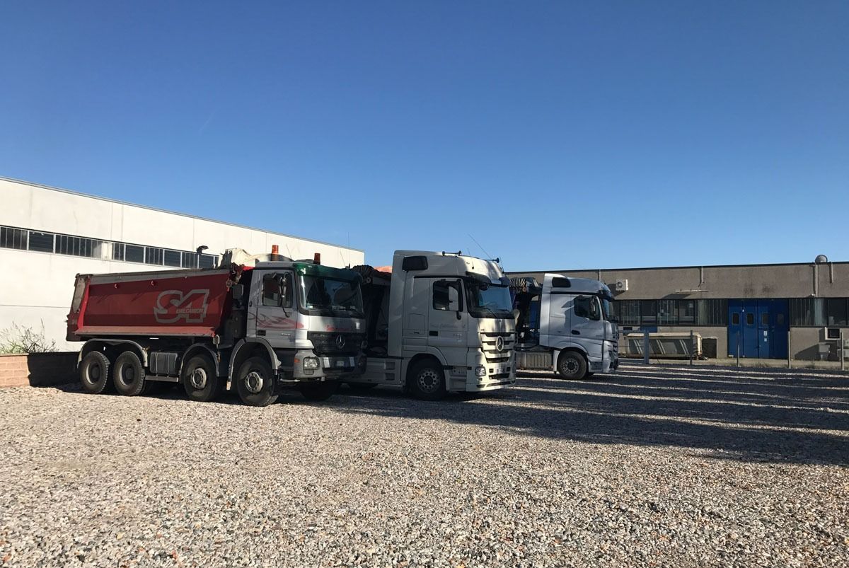 Tre camion parcheggiati sulla ghiaia davanti a un edificio bianco e grigio sotto un cielo azzurro.